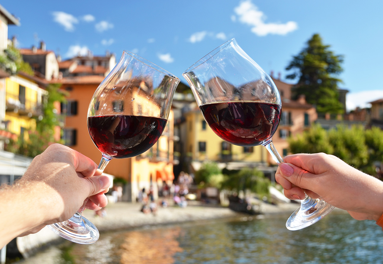 Two hands holding red wine glasses overlooking beautiful Varenna from the lake