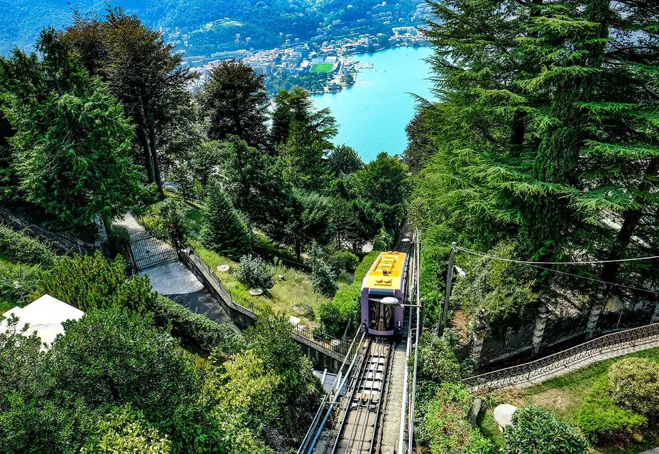 A  cable car ascending a mountain, surrounded by lush trees and a serene Como Lake