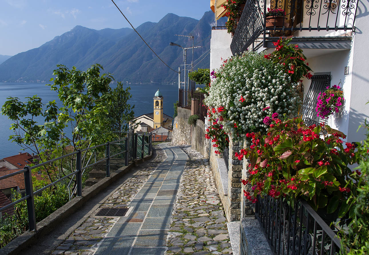 Ein gepflasterter Gehweg, der an weißen Häusern und bunten Blumen vorbeiführt und malerische Ausblicke auf einen malerischen See und eine kleine Kirche bietet