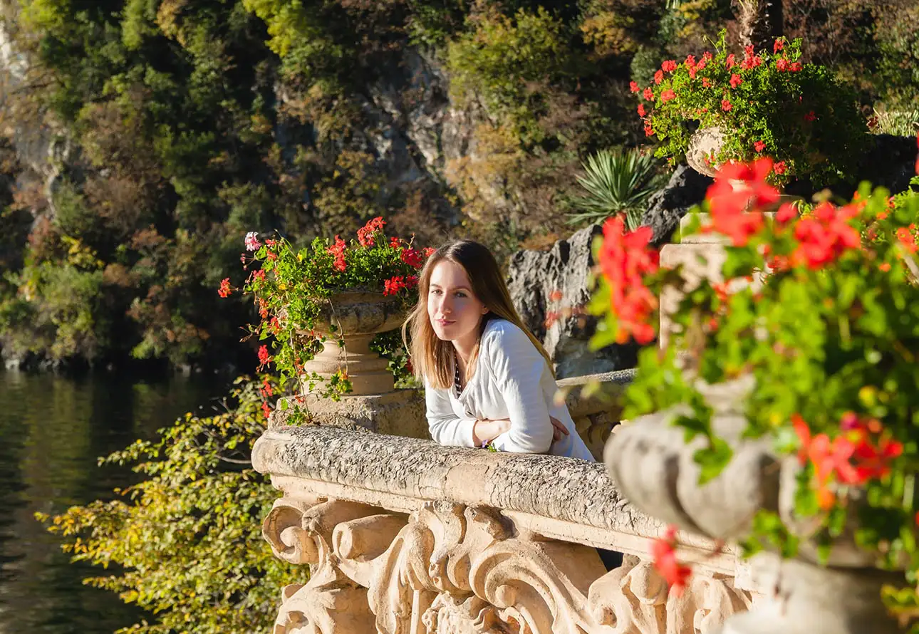 A woman gracefully leans from beautiful flowery balcony of Villa del Balbianello gazing out over the serene lake in a beautiful warm light.